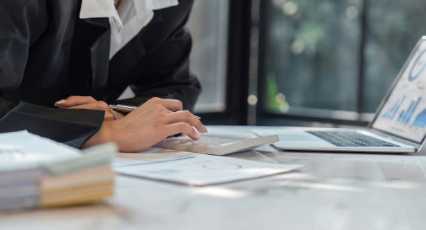 Business woman calculating monthly expenses, managing budget. Woman sitting at table using calculator to calculate tax refund, working at office with laptop on table.